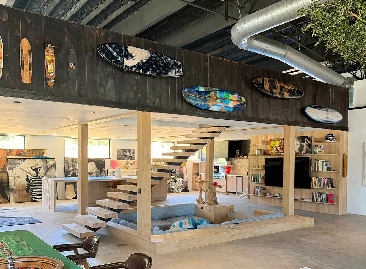 The studio interior in Kingston Springs — painted surfboards and skate decks racked along the dark beam above the sunken conversation pit, with the floating stair, the kitchen and the olive tree beyond