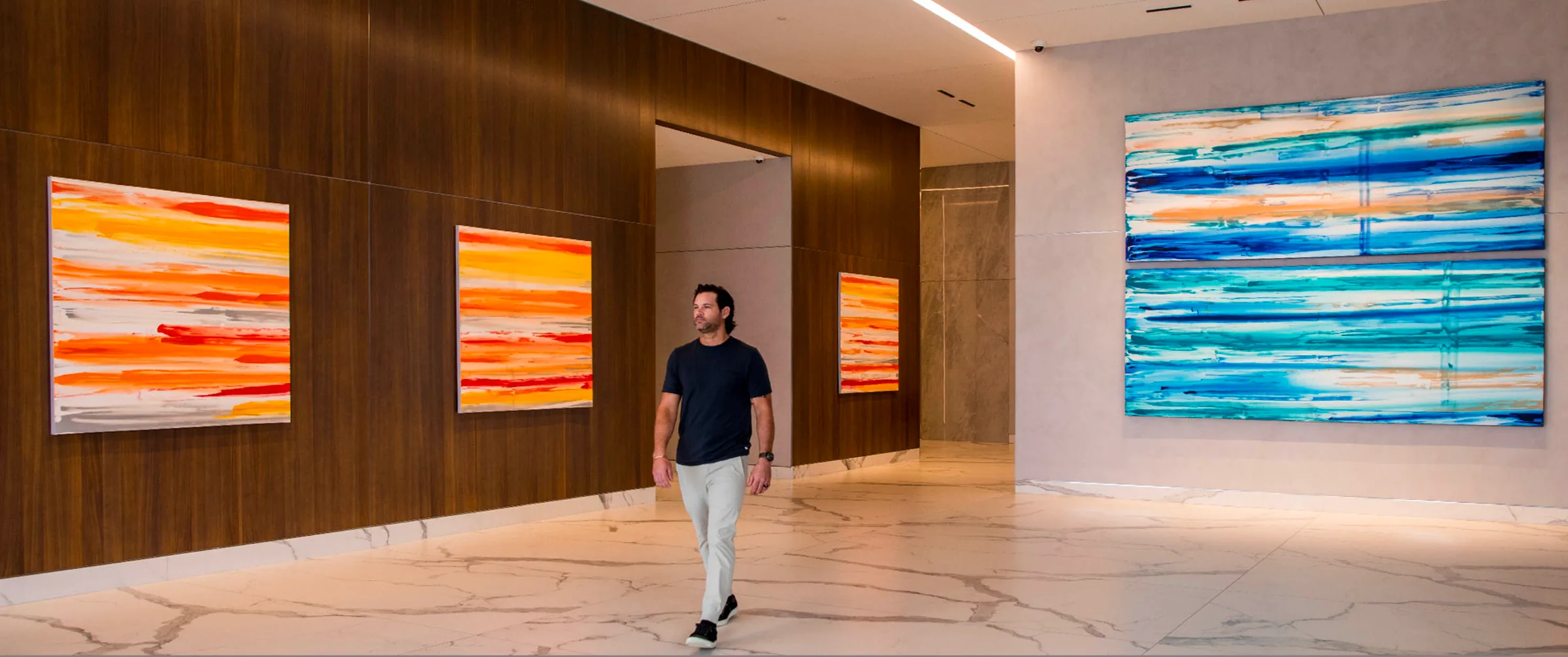 Erik Skoldberg walking through a wood and marble interior hung with three orange Urban Collection canvases on one wall and a large blue triptych on the other